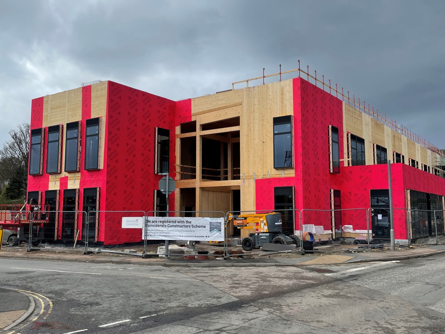 A photo of showing the progress of construction of a new office, family centre, and library at Union Street, Ellon. The photo shows the two-storey wooden frame of the building, with most of the windows installed. The site is bounded on two sides by roads, and protective metal fencing surrounds the site. Some construction equipment is within the site.  
