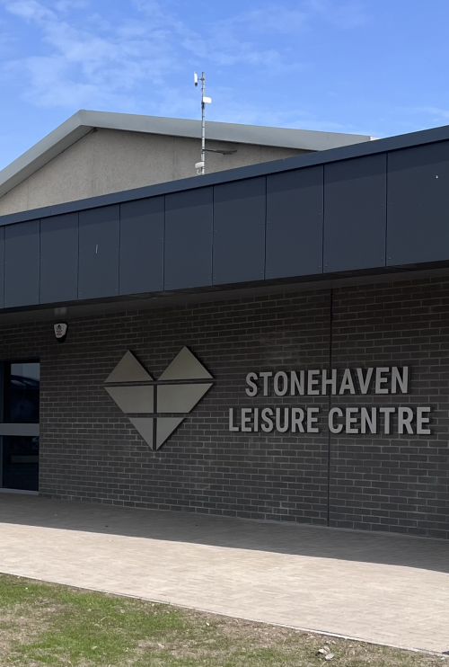 A photo of the front entrance of Stonehaven Leisure Centre, with a grey heart-shaped logo and Stonehaven Leisure Centre sign fixed to the wall. 