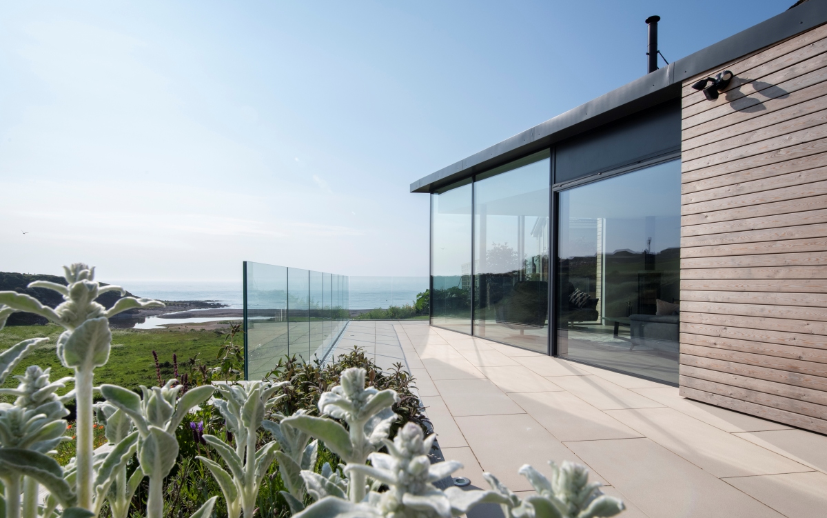Side of building with glass wall and glass balcony looking out at sea and plants in foreground