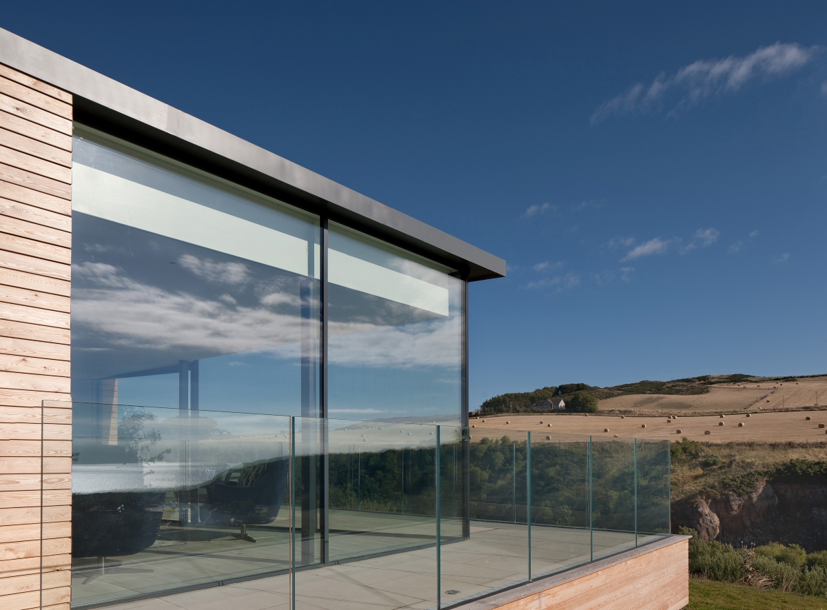 Side of building with glass wall and glass balcony looking out at hill with fields in background