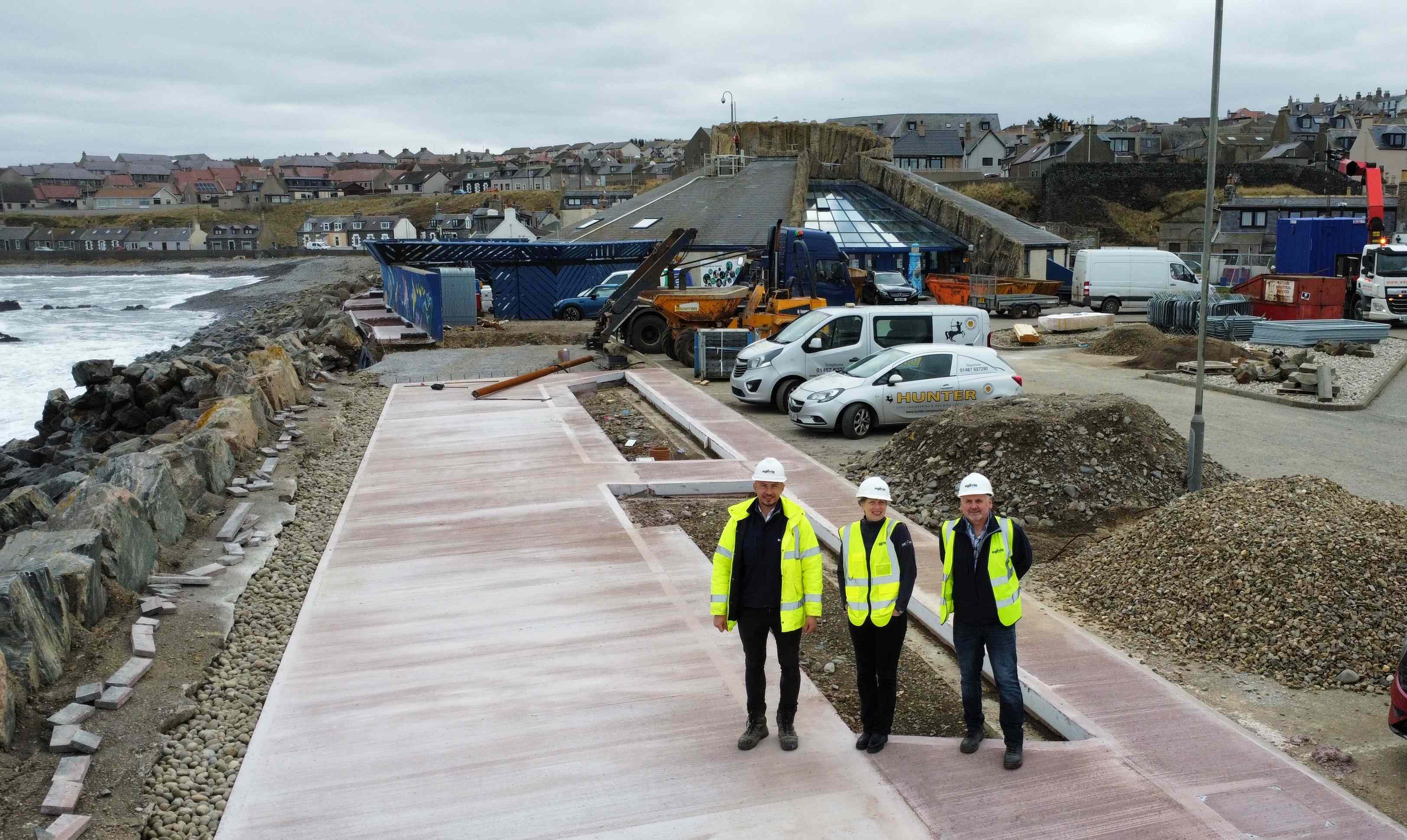 Three people pictured in high viz yellow vests and white hard hats in front of the Macduff Marine Aquarium with building materials on site   they are Ogilivie Construction project site manager main works Dave Brewis Macduff Marine Aquarium manager Claire Matthews and Ogilvie project site manager landscaping works Lucjan Binkowicz 