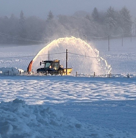 A photo of a vehicle clearing snow on a road between two fields. There is an arc of snow from a snow blower above the vehicle. 
