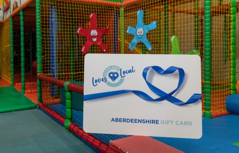 A colourful picture of an indoor children's play area with a large poster of the Aberdeenshire Loves Local gift card in white with blue writing