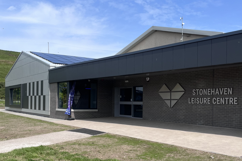 A photo of the main entrance of Stonehaven Leisure Centre, featuring a canopy that provides shelter in front of the entrance. A Stonehaven Leisure Centre sign and heart-shaped logo is fixed to the outer wall. 