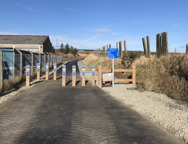 A picture showing a newly-resurfaced section of the Formartine and Buchan Way with wooden bollards to prevent vehicular access and a signed showing distances to villages and towns