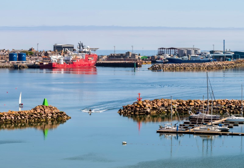 A colourful picture of Peterhead marina and bay featuring a red and white offshore deep sea support vessel, blue fishing boats and a jetski entering the marina