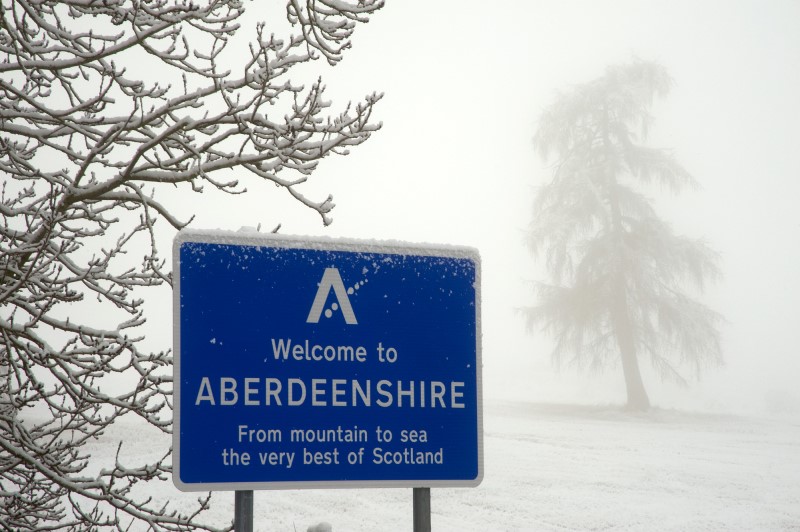 A wintry snow scene featuring a blue Welcome to Aberdeenshire road sign with white writing