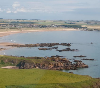 A photo of the Aberdeenshire coast, with a green field in the foreground leading to a rocky shore. A sandy beach is at the other side of the water, with a town in the background. 