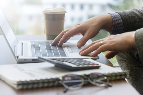 Close up of a laptop and hands typing with a coffee cup on one side and notebook with glasses on top on the other