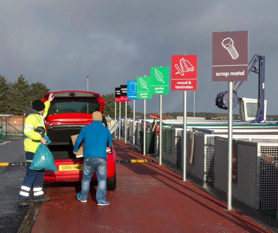 A household recycling centre with a visitor and staff member getting things out the back of a car.