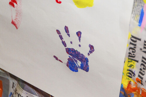 A photo of a child's handprint in purple, blue, and red paint on a white piece of paper. The paper is on top of paint-splattered newspaper. 