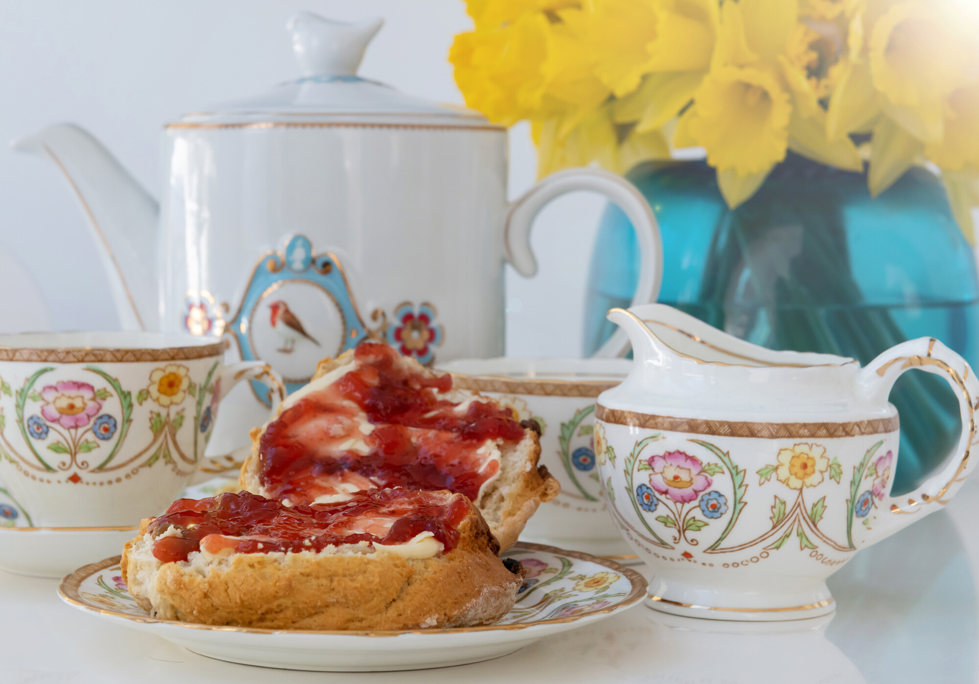 A picture of a tea set with floral pattern and a scone with cream and jam. There is a blue vase with daffodils in the rear of the picture