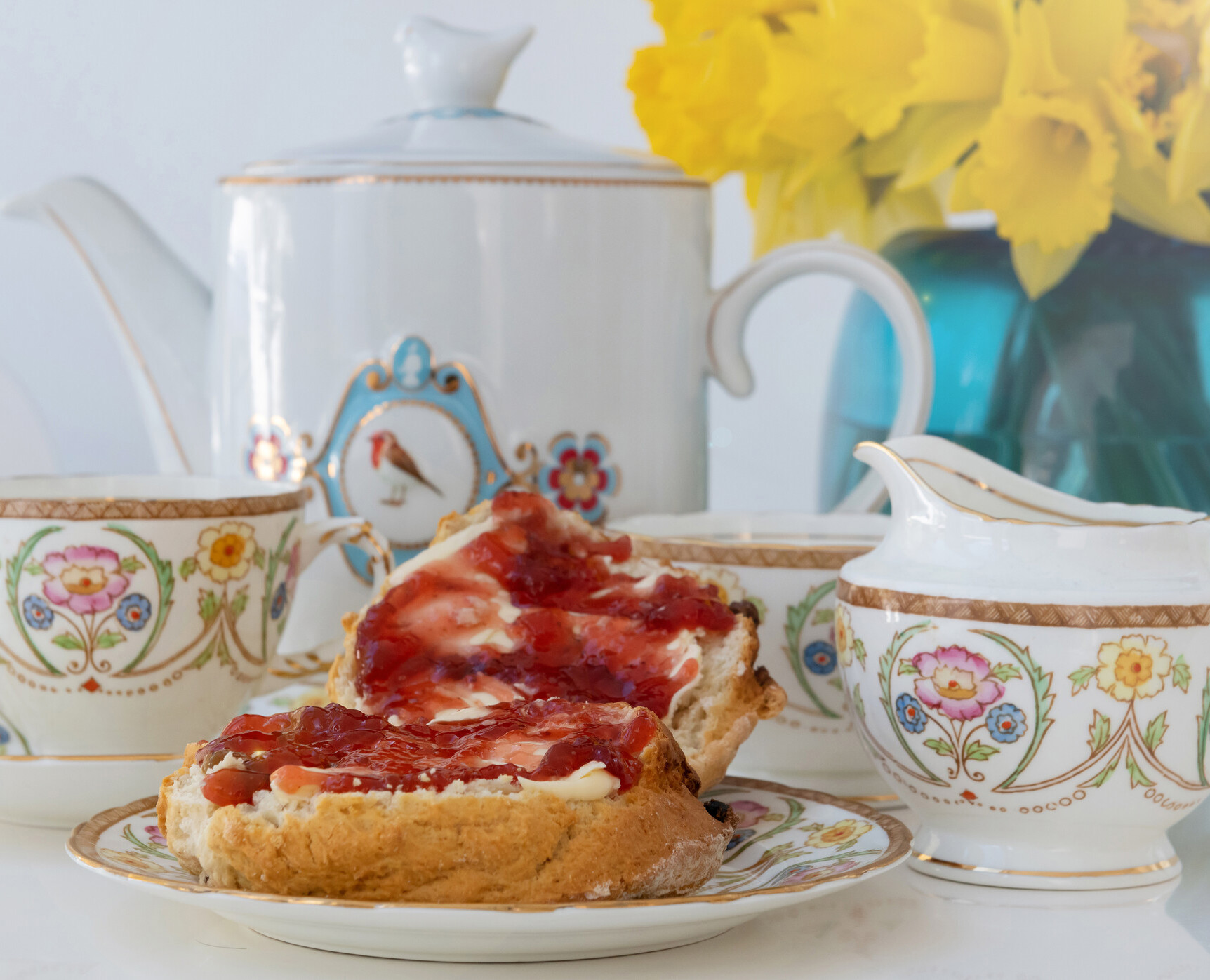 A picture of a tea set with floral pattern and a scone with cream and jam. There is a blue vase with daffodils in the rear of the picture